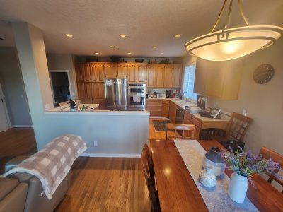 Open kitchen with natural wood cabinets, stainless steel fridge and double ovens, seen from a dining area in the foreground.