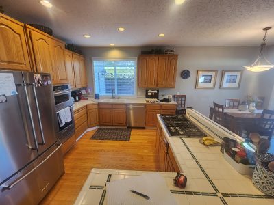 Bright kitchen with oak cabinets, stainless fridge, and a tiled island; dining table to the right and a window above the sink.