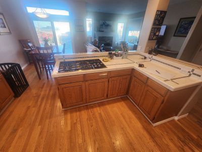 Open kitchen with white tiled counter and gas stove; view into living room with fireplace and a person seated on a sofa by the windows.