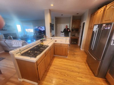 Man standing in doorway of a kitchen, waving hello, with an island, stove, and wooden cabinets visible; living room area and TV in background.