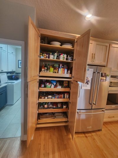 Open pantry cabinet in a kitchen, shelves stocked with cans, jars, bottles, and snacks; a stainless fridge is nearby.