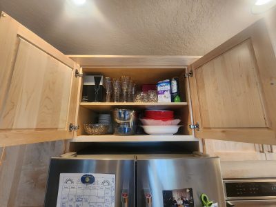 Open kitchen cabinet displaying wine glasses, bowls, and storage boxes; stainless steel fridge below with calendar and photos.