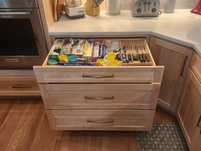 Open kitchen drawer filled with utensils, measuring cups, and a knife block organizer inside a wooden cabinet.