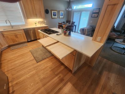 Open kitchen with light wood cabinets, marble countertops, stainless dishwasher, gas stove, and an island breakfast bar under warm lighting.