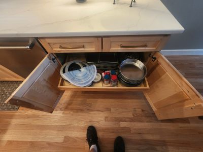 View from above of a kitchen island with a marble countertop, open drawers showing pots, pans, and spice jars inside. Feet visible in foreground.