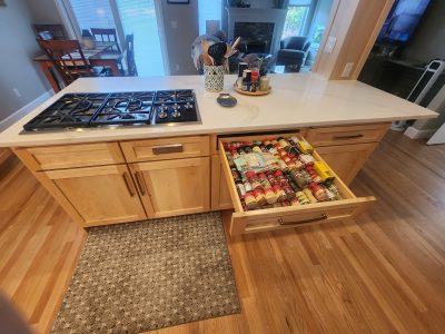 Kitchen island with a gas stove, spice drawer open, and utensil holder on a white countertop; dining area and living room visible beyond.