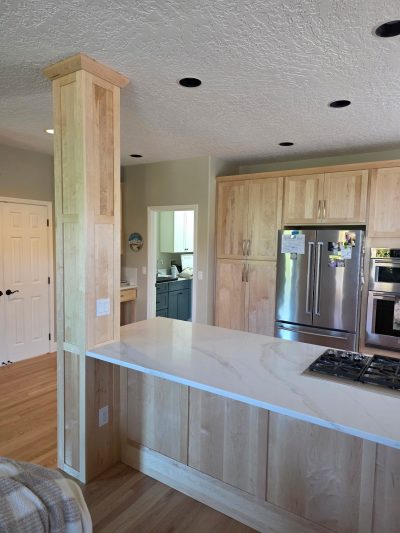 Kitchen with a marble island, light wood cabinets, and a stainless steel refrigerator; a large wooden column foregrounds the scene.